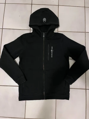 Svart zip hoodie från Sail Racing Bowman strl 160 - Svart zip hoodie från Sail Racing bowman (pojke strl 160) med huva och hel dragkedja framtill. Hoodien har en bröstficka med vertikal dragkedja och Sail Racing-logga tryckt på bröstet. Snygg, stilren design med långa ärmar och ribbade muddar. Perfekt för blåsiga dagar. 