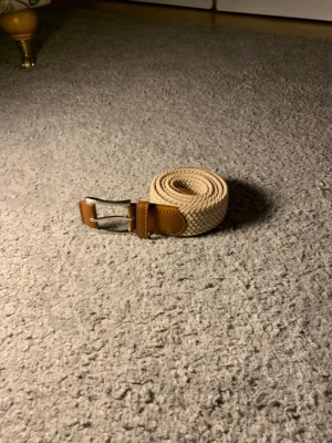 Beige flätat bälte med läderdetaljer - Snyggt beige flätat bälte med bruna läderdetaljer och silverfärgat spänne. Perfekt accessoar för att lyfta din outfit med en stilren och modern touch. Bältet är flexibelt och passar till både jeans och chinos.