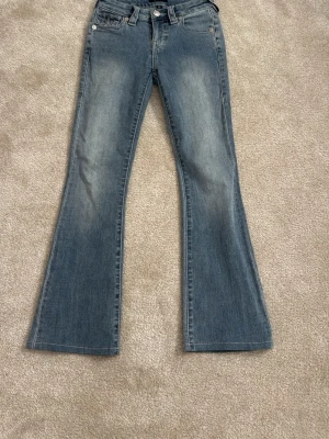 TRUE RELIGION Blå bootcut jeans med fickdetaljer s - Snygga blå bootcut jeans med klassisk femficksdesign och dekorativa ficklock bak. Jeansen har en lätt utsvängd passform och subtila slitningar framtill för en sopad vintagekänsla. Perfekta till sneakers eller boots.