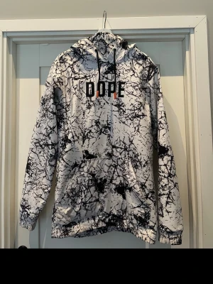DOPE hoodie med svartvitt mönster - Säljer en hoodie från DOPE med ett unikt svartvitt marmorerat mönster och svart logga på bröstet. Hoodien har huva med svarta snören och magficka framtill. Perfekt för dig som vill sticka ut med streetwear-stil.