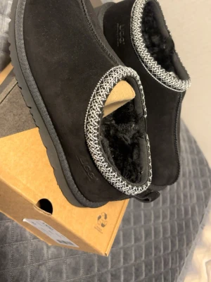 Svarta UGG tofflor i mocka - Mysiga svarta tofflor från UGG i mjuk mocka med fluffigt foder och dekorativt mönster runt öppningen. Perfekta för att hålla fötterna varma hemma. Klassisk UGG-logga på sidan och rejäl sula för extra komfort.