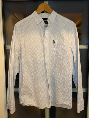 Ljusblå randig skjorta Lyle & Scott - Säljer en ljusblå och vit prick, randig skjorta från Lyle & Scott med klassisk krage och lång ärm. Skjortan har en bröstficka. Tillverkad i ett strukturerat bomullstyg som ger en avslappnad men stilren look. Strl: M vid funderingar fråga gärna!🛥️