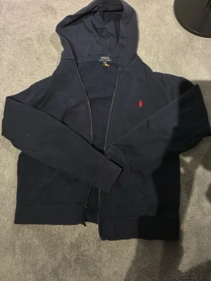 Mörkblå hoodie från Polo Ralph Lauren - Snygg mörkblå hoodie med dragkedja från Polo Ralph Lauren. Klassisk design med huva, fickor framtill och den ikoniska röda loggan broderad på bröstet. Perfekt för en avslappnad och stilren look.