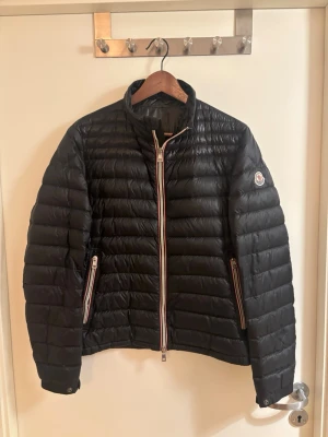 Moncler Daniel Short Down  - Snygg svart dunjacka från Moncler med klassisk quiltad design och raka linjer. Jackan har dragkedja framtill med Monclers signaturfärger och två fickor med dragkedja. Liten Moncler-logga på ärmen. Perfekt för kyliga dagar. Köpt från moncler hemsida några säsonger sedan. Varken kvitto eller box förekommer. Nypris allt från 11.000-13.000kr. 
