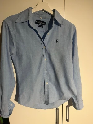 Ljusblå skjorta från Ralph Lauren - Klassisk ljusblå skjorta från Ralph Lauren i 100% bomull. Skjortan har lång ärm, knappar framtill och den ikoniska broderade loggan på bröstet. Perfekt till jeans eller chinos för en clean och stilren look.