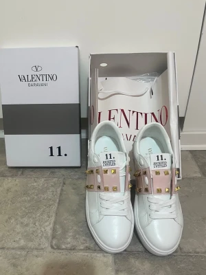 Valentino Rockstud sneakers vit/rosa - Säljer ett par Valentino Garavani Rockstud sneakers i vitt skinn med ljusrosa detaljer och guldiga nitar. Klassisk rund tå, snörning och platt sula. Ikoniska pyramidnitar längs sidorna och bak på hälen. Kommer med originalkartong och dustbag. Passar dig som vill ha en lyxig stil nu i det nya året.
