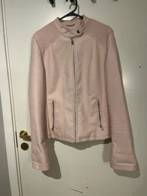 Ljusrosa skinnjacka med dragkedjor - Snygg ljusrosa skinnjacka med ståkrage och silverfärgade dragkedjor framtill och vid fickorna. Jackan har ribbade detaljer på axlar och sidor samt dragkedjor vid ärmsluten. Perfekt för dig som vill ha en stilren och trendig look. Storleken är XXL men sitter som en S