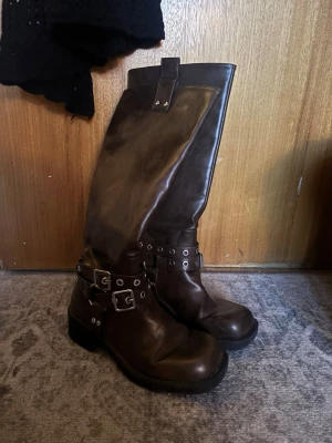 Bruna höga läderstövlar med spännen - Snygga mörkbruna stövlar i skinn med högt skaft och coola metallspännen runt ankeln. Rund tå och rejäl platt sula med ca. 2 cm klack. Använda fåtal gånger.
