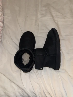 Svarta UGG boots i mocka - Klassiska svarta UGG boots i mjuk mocka med fårskinnsfoder. Modellen har rund tå, platt sula och en enkel design utan snörning. Perfekta för kalla dagar och ger en mysig känsla. UGG-logga bak på hälen.