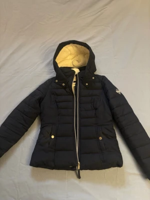 Svart dunjacka med huva från Hollister - Snygg svart puffer jacket från Hollister med mysigt teddyfoder på insidan och i huvan. Jackan har dragkedja framtill, två fickor med tryckknappar och en klassisk passform. Perfekt för vår/höst dagar.