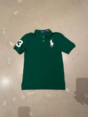 Grön pikétröja från Polo Ralph Lauren - Klassisk grön pikétröja från Polo Ralph Lauren med vit logga på bröstet och siffran 3 på ärmen. Tröjan har krage, knappar framtill och är kortärmad. Classic fit-modell som passar till mycket. Perfekt för dig som gillar sportig och stilren look.