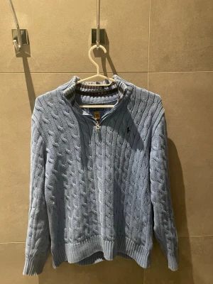Ljusblå kabelstickad tröja Polo Ralph Lauren - Ljusblå kabelstickad tröja från Polo Ralph Lauren med halv dragkedja och ribbad krage. Tröjan har långa ärmar, ribbade muddar och den klassiska broderade loggan på bröstet. Perfekt för lager på lager och en chill look. Strl 14-16