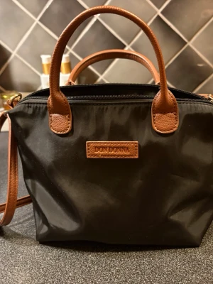 Svart handväska från Don Donna - Snygg svart handväska från Don Donna med bruna detaljer i skinnimitation. Väskan har både handtag och axelrem samt dragkedja upptill. Perfekt storlek för det viktigaste och enkel att matcha till olika outfits.