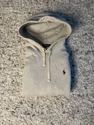 Grå hoodie från Polo Ralph Lauren - Snygg ljusgrå hoodie från Polo Ralph Lauren med klassisk broderad logga på bröstet. Hoodien har dragsko i huvan och är gjord i mjukt bomullsmaterial. Det finns en defekt som visas på tredje bilden.