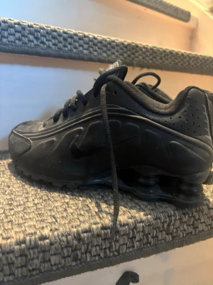 Svarta Nike Shox sneakers - Nike Shox sneakers i helsvart med ikoniska fjädrande pelare i sulan. Skorna har snörning, rund tå och är gjorda i syntetmaterial med meshdetaljer. Swoosh-logga på sidan och Nike Shox-tryck på plösen. Perfekt för dig som gillar sportig och futuristisk stil. Jag har inte orginal lådan eller kvittot för skorna är ganska gammal men är i väldigt bra skick