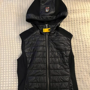 Parajumpers väst - Snygg svart dunväst från Parajumpers med huva och gult spänne vid halsen. Västen har quiltad framsida, dragkedja och broderad logga på huvan. Perfekt lager-på-lager-plagg för kyliga dagar. Sportig och stilren look. Har använt den bara en gång och om du är intresserad så kan jag skicka qr koden så att du vet att den är äkta.