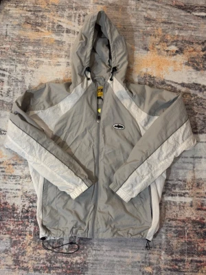 Grå Corteiz vindjacka med huva - Snygg grå vindjacka från Corteiz med vita detaljer och hel dragkedja framtill. Jackan har huva med snörning, elastiska muddar och justerbar nederkant. Perfekt för blåsiga dagar och enkel att matcha till olika outfits.