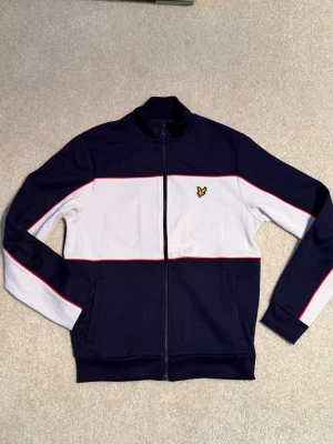 Marinblå retro ziptröja från Lyle & Scott - Snygg marinblå ziptröja från Lyle & Scott med vit panel och röda detaljer på ärmarna. Tröjan har ståkrage, hel dragkedja och två fickor framtill. Ikonisk gul logga på bröstet. Perfekt för en sportig och stilren look.
