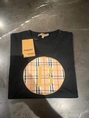 Svart Burberry t-shirt med logga - Svart t-shirt från Burberry med klassiskt rutigt märke och stor gul B-logga på bröstet. T-shirten har rund halsringning och korta ärmar. Perfekt för dig som gillar stilrena plagg med exklusiv känsla.