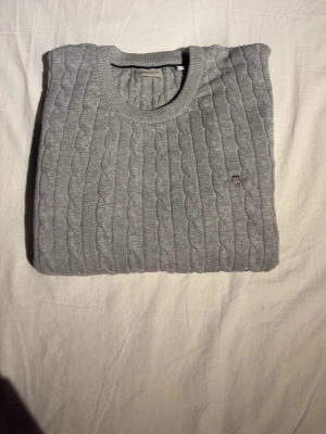 Grå kabelstickad tröja från Gant - Snygg grå kabelstickad tröja från Gant med rund halsringning och diskret logga på bröstet. Tröjan har långa ärmar och ribbade muddar vid ärmslut och nederkant. Perfekt för lager på lager och enkel att matcha med jeans eller kjol.