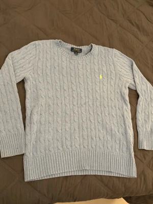 Ljusblå kabelstickad tröja Polo Ralph Lauren - Ljusblå kabelstickad tröja från Polo Ralph Lauren med rund halsringning och klassisk gul broderad logga på bröstet. Tröjan har långa ärmar och ribbade muddar vid ärmslut och nederkant. Perfekt för en stilren och avslappnad look.