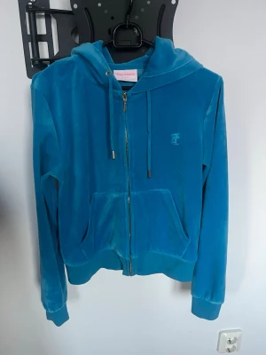 Blå velourhoodie från Juicy Couture - Snygg blå (åt det turkosa hållet) velourhoodie från Juicy Couture med guldfärgad dragkedja, huva och fickor framtill. Klassisk JC-logga broderad på bröstet och guldfärgade detaljer på snörena. Något liten i storleken.