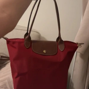 Röd handväska från Longchamp - Snygg röd handväska från Longchamp med bruna skinnhandtag och lock. Väskan har klassisk form, guldfärgade detaljer och dragkedja upptill. Perfekt för dig som vill ha en stilren och rymlig väska till vardagen.
