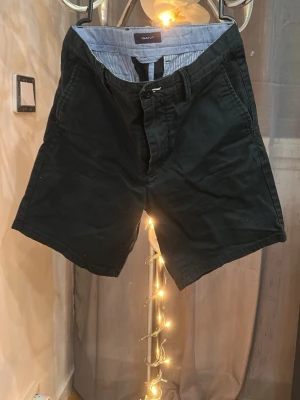 Svarta shorts från GANT - Klassiska svarta shorts från GANT med normal passform. Tillverkade i bomull med fickor på sidorna och knappstängning framtill. Insidan har ett ljusblått och vitt randigt foder som ger en snygg detalj.