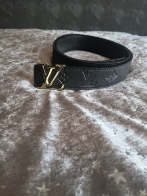 Svart Louis Vuitton bälte i läder - Snyggt svart bälte från Louis Vuitton i präglat läder med klassiskt LV-mönster och en guldig LV-logga som spänne. Perfekt accessoar för att lyfta din outfit med en lyxig touch.