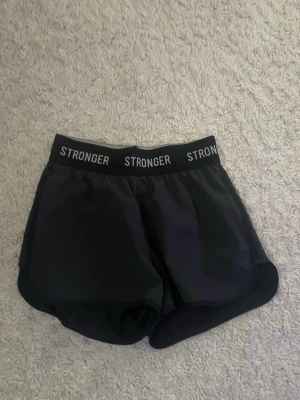 Svarta träningsshorts från Stronger - Snygga svarta träningsshorts från Stronger med elastisk midja där loggan är tryckt runtom. Shortsen är i lätt och snabbtorkande material med meshfoder på insidan. Perfekta för löpning, gym eller annan träning.