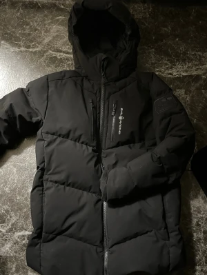 Sail racing vinterjacka  - Snygg svart dunjacka från Sail Racing med huva och dragkedja framtill. Jackan har en quiltad design, bröstficka med dragkedja och logga på bröstet. Perfekt för kalla dagar och enkel att matcha med allt.
