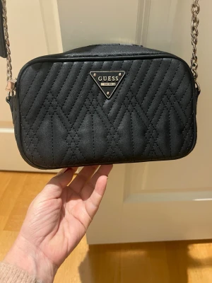 Svart quiltad handväska från Guess - Snygg svart quiltad handväska från Guess med kedjerem och triangelformat metallogo framtill. Väskan har ett stilrent mönster och är tillverkad i syntetmaterial. Perfekt för dig som vill ha en trendig accessoar med plats för det viktigaste.