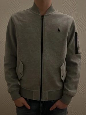 Grå kofta med dragkedja - Snygg grå kofta från Polo Ralph Lauren med dragkedja framtill, ribbade muddar och fickor med lock. Koftan har en extra ficka med dragkedja på ärmen. Perfekt för en stilren och avslappnad look. Aldrig använd, inga tecken på användning. Storlek S. Hör av er vid eventuella frågor