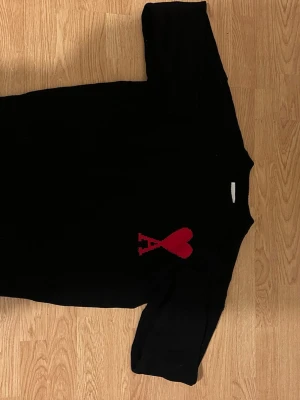 Svart stickad tröja från Ami - Snygg svart stickad tröja från Ami med röd hjärtlogga och bokstaven A på bröstet. Tröjan har rund hals och ribbade muddar. Perfekt för dig som gillar stilrena plagg med en twist. pris kan diskuteras 