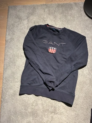 Mörkblå sweatshirt från GANT - Säljer en mörkblå sweatshirt från GANT med broderad logga och sköld på bröstet. Tröjan har rund halsringning, långa ärmar och ribbade muddar. Perfekt för en avslappnad och stilren look.