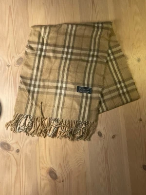 Beige rutig halsduk från Burberry - Klassisk beige halsduk från Burberry med rutigt mönster i brunt, vitt och svart. Halsduken har fransar i ändarna och en vävd Burberry-logga. Perfekt accessoar för att lyfta din outfit. Pris kan diskuteras.