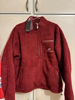 Vinröd teddytröja från nohalfsends - Mysig vinröd teddytröja från nohalfsends med halv dragkedja framtill, bröstficka med dragkedja och broderad logga på bröstet samt stor brodyr på ryggen. Tillverkad i fluffigt teddy-material som håller dig varm och skön.