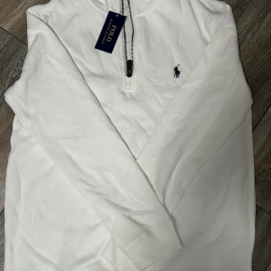 Vit half zip tröja från Polo Ralph Lauren - Vit långärmad tröja från Polo Ralph Lauren med half zip och klassisk broderad logga i marinblått på bröstet. Tröjan är i mjuk bomull och har ribbade muddar vid ärmslut och hals. Perfekt för en clean och stilren look.