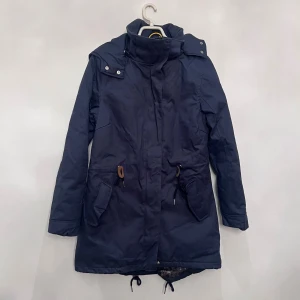 Mörkblå parkajacka  - Parkajacka i mörkblått från Lyle & Scott med huva, högt krage och snörning i midjan. Jackan har quiltat foder, dragkedja och knappar framtill samt två stora fickor. Perfekt för kalla dagar och har en clean, urban vibe.