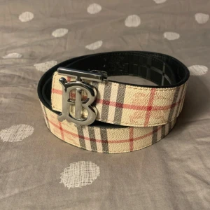 Burberry vändbart skärp med B-spänne - Snyggt vändbart bälte från Burberry med klassiskt rutigt mönster i beige, svart och rött på ena sidan och svart präglat skinn med diskret logga på andra. Stort silvrigt B-spänne framtill. Perfekt accessoar för att lyfta din outfit.