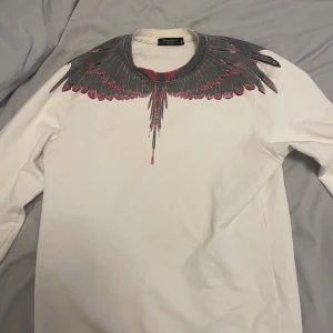 Vit sweatshirt Marcelo Burlon med vingar - Säljer en vit långärmad sweatshirt från Marcelo Burlon med ikoniskt vingmönster i svart och rött runt halsen och axlarna. Tröjan har rund hals och ribbade muddar. Perfekt för dig som vill sticka ut med en unik design.