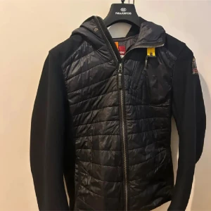 Svart quiltad jacka från Parajumpers - Svart jacka från Parajumpers med quiltad front och rygg, huva och dragkedja. Jackan har en gul detalj vid halsen och en patch på ärmen. Materialmix med vadderad kropp och släta ärmar ger en sportig vibe.