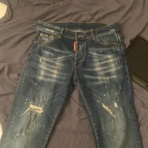 Dsquared2 blå distressed jeans - Snygga blå jeans från Dsquared2 med slitna detaljer, färgstänk och trasiga partier på benen. Klassisk femficksmodell med röd Dsquared2-tag vid gylfen och patch bak. Jeansen har en smal passform och cool tvättad look.