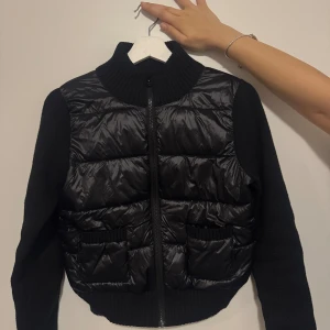 Svart pufferjacka med stickade ärmar - Svart pufferjacka med stickade ärmar och rygg, samt ribbad hög krage. Framsidan har quiltad pufferpanel i blankt material och två fickor med ribbade detaljer. Dragkedja framtill och tight passform. Perfekt för dig som gillar en clean och modern look. Storleken är M men själv är Xs-S men eftersom denna är lite tajt då M passar perfekt för dig också som har en storlek  Xs eller S. Har använt den bara 2 gånger.