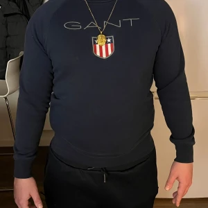 Mörkblå sweatshirt från GANT - Snygg mörkblå sweatshirt från GANT med broderad logga och emblem på bröstet. Tröjan har rund hals, långa ärmar.Perfekt för dig som gillar klassisk stil med sportig touch. Materialet är mjukt och skönt, troligen bomull och polyester.