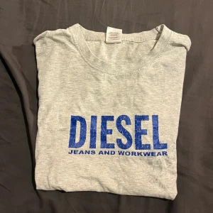 Grå t-shirt från Diesel med blå tryck - Snygg grå t-shirt från Diesel med stort blått tryck på bröstet där det står 'DIESEL JEANS AND WORKWEAR'. Klassisk rund hals och korta ärmar. Perfekt för en avslappnad streetstyle-look.
