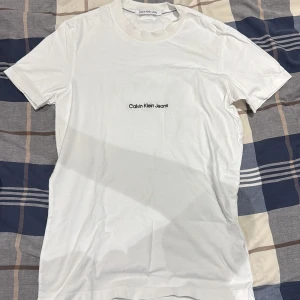 Vit t-shirt från Calvin Klein Jeans - Snygg vit t-shirt från Calvin Klein Jeans med svart logotyp tryckt på bröstet och diskret CK-broderi i nacken. Klassisk rund hals och korta ärmar. Mjuk och skön bomullskvalitet som känns fräsch och enkel att matcha.