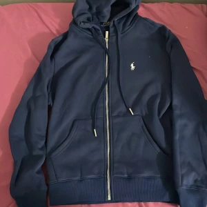 Marinblå hoodie från Polo Ralph Lauren - Snygg marinblå hoodie från Polo Ralph Lauren med klassisk vit broderad logga på bröstet. Tröjan har huva med snörning, dragkedja framtill och rymliga fickor. Mjuk insida i fleece och ribbade muddar vid ärmslut och nederkant. Perfekt för chill dagar.