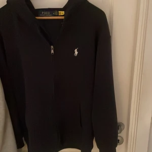Svart hoodie från Polo Ralph Lauren - Svart hoodie från Polo Ralph Lauren med klassisk vit logga på bröstet. Tröjan har dragkedja framtill, huva och långa ärmar. Materialet är mjukt och känns som bomull, perfekt för chill dagar. Snygg och enkel design som funkar till det mesta.