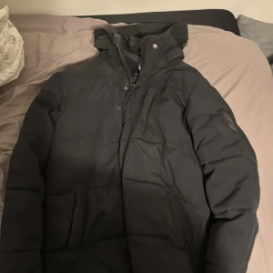 Svart pufferjacka från five seasons - Säljer en svart pufferjacka med huva och hög krage. Jackan har dragkedja och tryckknappar framtill samt praktiska fickor på sidorna. Perfekt för kalla dagar och har en stilren, enkel design som passar till det mesta.